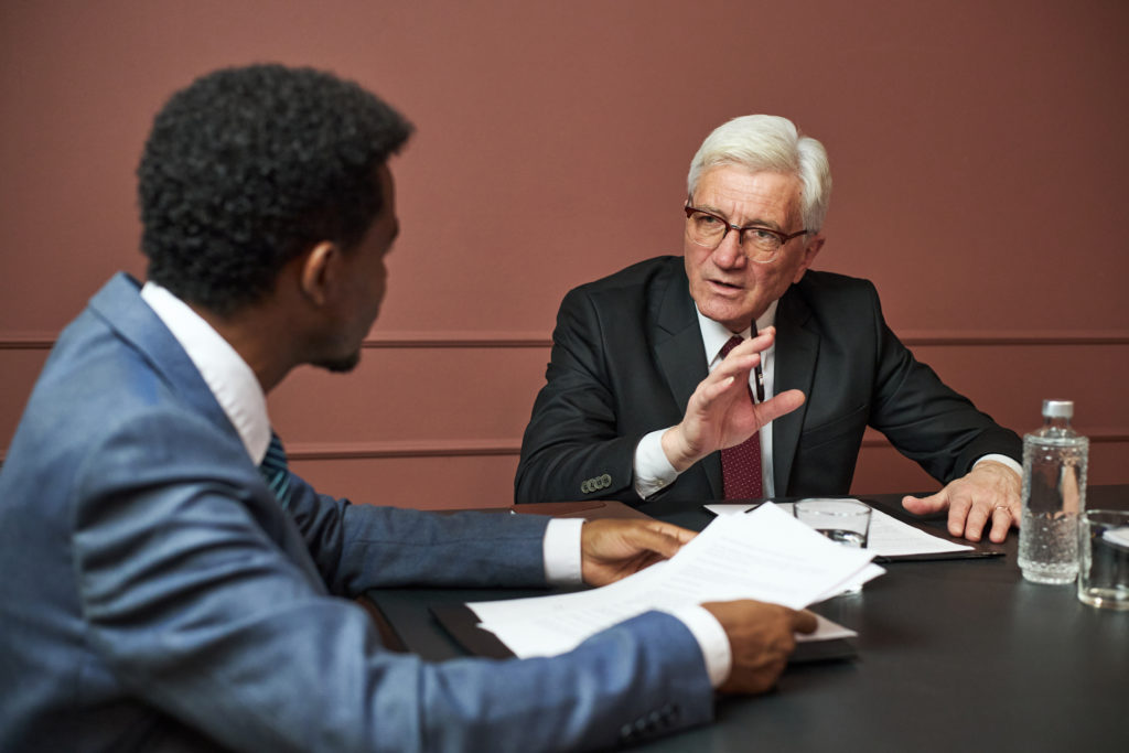Middle aged Black man and senior Caucasian man discussing documents at table, senior man gesturing while speaking, both wearing business attire, water bottle and papers visible